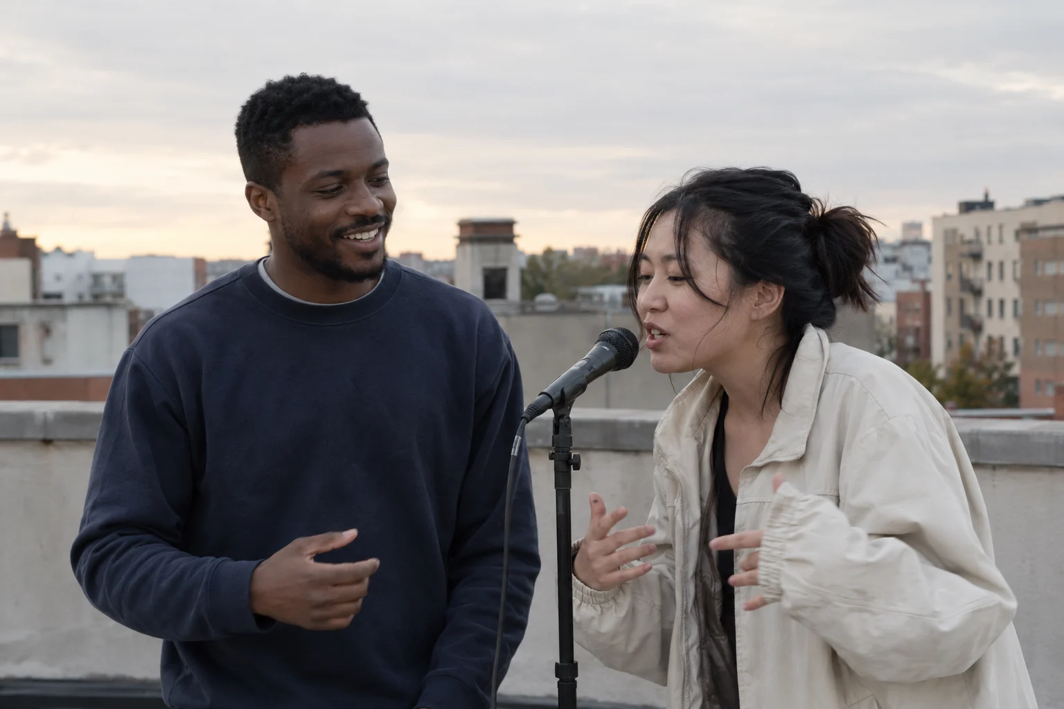 Two fictional friends sharing a microphone on a rooftop in soft evening daylight
